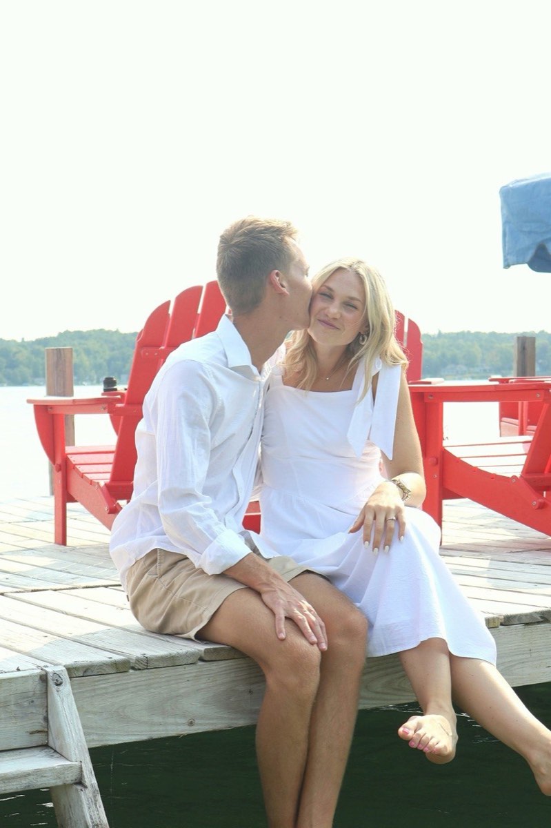 Tucker kissing Syd on the cheek as they sit on a dock